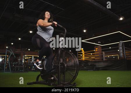 Cardio, weight loss, body positive concept. Low angle shot of a cheerful plus size sportswoman exercising on air bike at sport studio. Curly haired ov Stock Photo