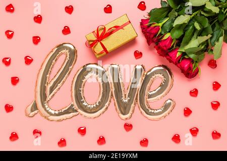 Love word shaped with shiny foil balloon, red roses bouquet, golden gift box and wooden hearts on pink background. Mothers, womens or Valentines day Stock Photo