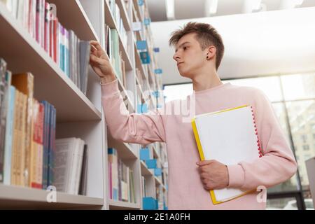 Low angle shot of a male student taking a book from the shelf at college library. Young male customer shopping for books at the bookstore Stock Photo
