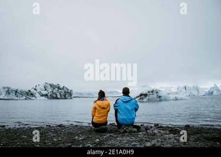 Unrecognizable couple sitting their backs. Looking at a glacier in Iceland. Yellow and blue coats. Concept of travel and love Space for text. Stock Photo