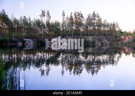 View of a beautiful clear lake among granite rocks and pine forest reflected in the water at sunset in summer Stock Photo