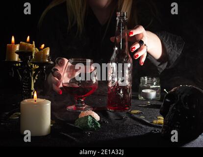 The witch's hand holds a bottle with a red liquid in the shape of a skull and a glass. Magical potion. The concept of wizards, witchcraft and magic. Stock Photo