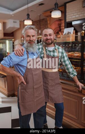 Vertical shot of a senior baker and his son smiling to the camera posing proudly at their family bakery store Stock Photo