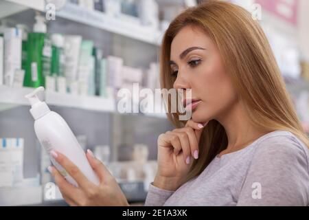 Close up of a beautiful woman rubbing her chin thoughtfully, reading label on a medication bottle at the pharmacy. Attractive female customer shopping Stock Photo