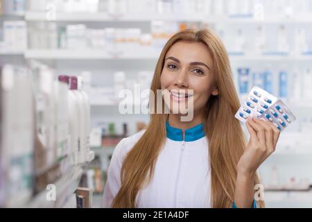 Close up of a cheerful professional pharmacist smiling joyfully, holding capsule package, posing at the drugstore. Attractive female pharmacist offeri Stock Photo