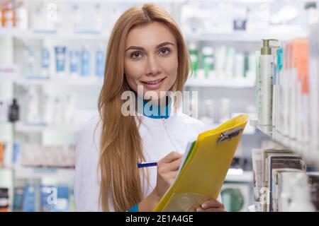 Lovely female pharmacist smiling to the camera, taking notes on her clipboard, working at the drugstore. Beautiful happy confident chemist checking st Stock Photo