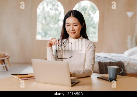 Young Asian businesswoman with glasses and laptop smiling at looking at camera. Female freelancer sitting at table and working from home. Stock Photo