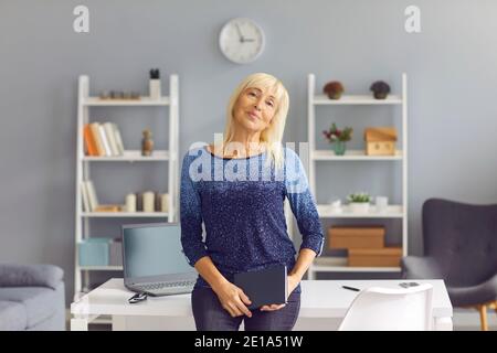 Portrait of senior business woman in casual clothes standing in office with notebook in hands. Stock Photo