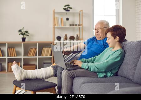 Happy supportive grandson and his granddad with broken leg using laptop together Stock Photo