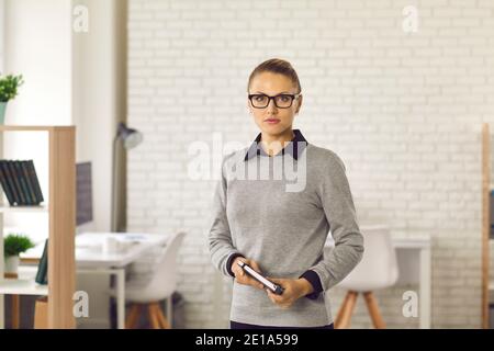 Serious woman in glasses and formal wear holding notebook and standing in office Stock Photo