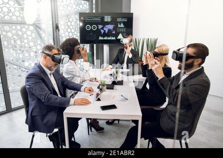 Focused five multiethnic professional business colleagues in formal wear, sitting at the table in office and using VR headsets. Business, technology Stock Photo