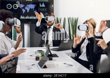 Business team of five multiracial diverse software engeneers, using virtual reality simulators, sitting at the table in office meeting, touching air Stock Photo