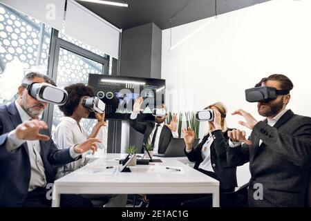 Business team of five multiracial diverse software engeneers, using virtual reality simulators, sitting at the table in office meeting, touching air Stock Photo