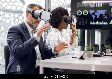 Two business persons, mature Caucasian man and young African woman, wearing vr goggles, developing a project or testing new mobile applications, while Stock Photo