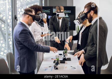 Team of creative multiracial colleagues, designers, architect in formal wear, standing around the table and using vr goggles for visualization of the Stock Photo