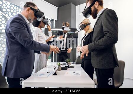 Team of creative multiracial colleagues, designers, architect in formal wear, standing around the table and using vr goggles for visualization of the Stock Photo