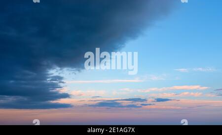 Large dark storm cloud obscures the clear pink blue vanilla sky at dawn. Calm before the storm, serenity after hurricane, light and dark confrontation Stock Photo