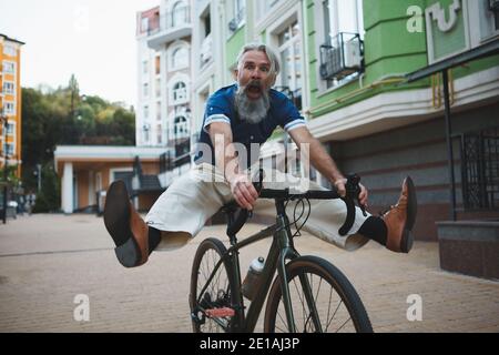 Senior bearded man looking funny, riding bicycle, screaming lifting his legs in the air Stock Photo