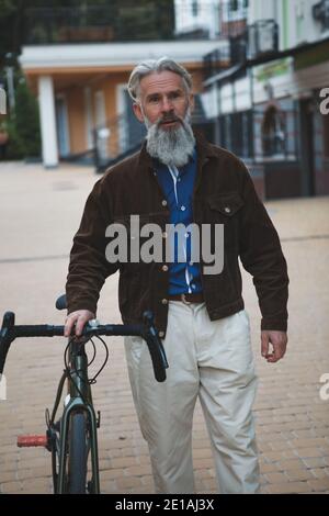 Vertical portrait of a bearded stylish mature man wearing fashionable jacket, walking with his bicycle Stock Photo