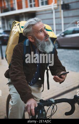 Vertical shot of a senior bearded man working as delicery guy, riding his bicycle, using smart phone Stock Photo
