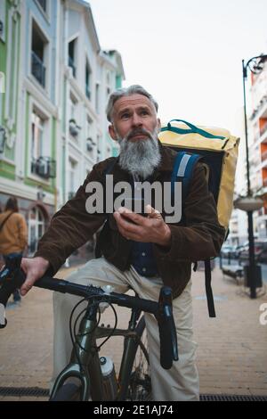Vertical portrait of a senior bearded man working as deliveryman on a bicycle, using smart phone in the city Stock Photo