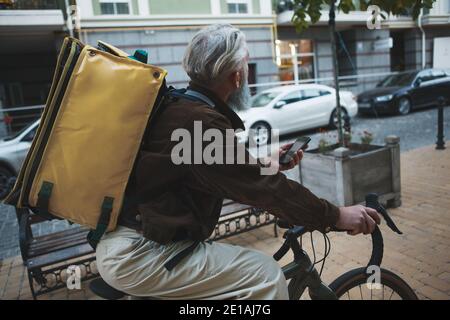 Rear view shot of a senior deliveryman riding bicycle, wearing delivery backpack Stock Photo
