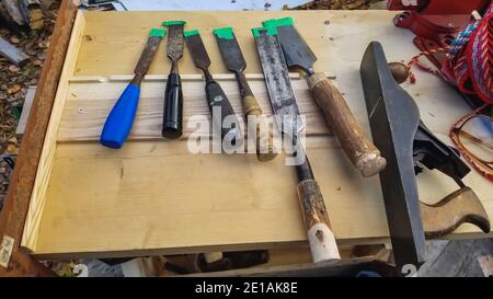 Wood chisels in variety of sizes placed together on a crafted light-colored wooden structure, other cutting tool and rope can be seen right next to them.  Stock Photo