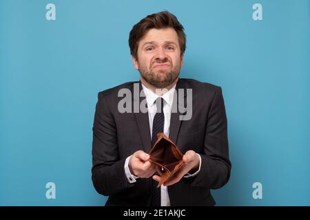 Businessman showing empty wallet having no money. I lost all my savings. Studio shot on blue wall. Stock Photo