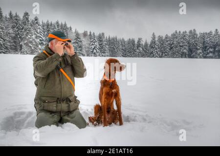 In winter a hunter kneels in the snow and observes his snowy hunting ground through a small eyepiece, next to him sits his Irish Setter dog. Stock Photo