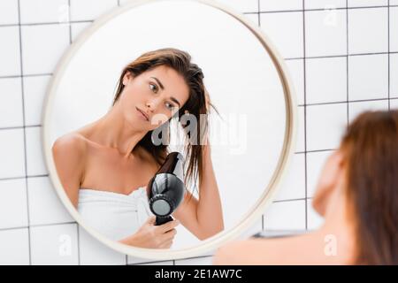 young woman looking in mirror while drying hair in bathroom on blurred foreground Stock Photo