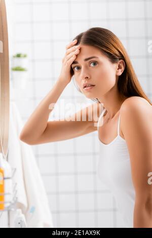 dissatisfied young woman looking at camera while touching forehead in bathroom Stock Photo