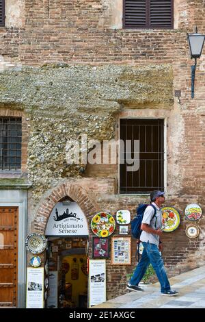 A tourist with a backpack walks in front of a medieval building with a souvenir shop in the historic centre of Siena, Unesco W.H. Site, Tuscany, Italy Stock Photo