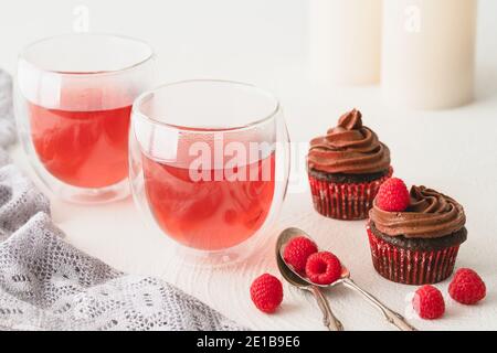Raspberry tea in glass cups and chocolate cupcakes decorated with chocolate frosting and fresh raspberries close up on white background. Sweet delicio Stock Photo