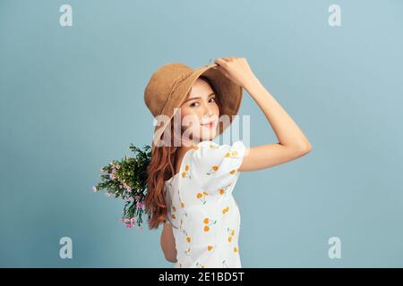 Close up portrait of an attractive young woman in summer dress and straw hat holding flower bouquet and looking over her shoulder isolated over blue b Stock Photo