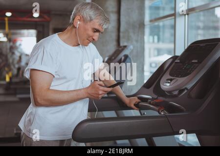 Senior healthy man listening to music on his smart phone, preparing for cardio workout on a treadmill. Elderly cheerful sportsman using smart phone wh Stock Photo