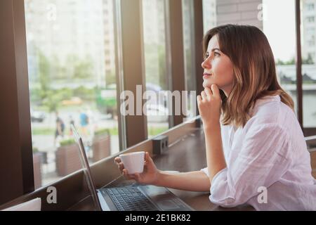 Attractive young businesswoman looking away thoughtfully, working on her laptop at the coffee shop. Beautiful woman working on a project on computer a Stock Photo