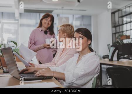 Group of successful businesswomen working at the office together. Lovely young businesswoman smiling to the camera, her female colleagues talking on t Stock Photo