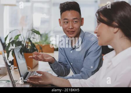 Cheerful Asian businessman talking to his female colleague at the office. Young businesspeople enjoying working on a creative project. Male designer t Stock Photo