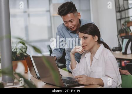 Young Asian businessman and his female colleague using laptop at work together. Attractive businesswoman working with her colleague, browsing online o Stock Photo
