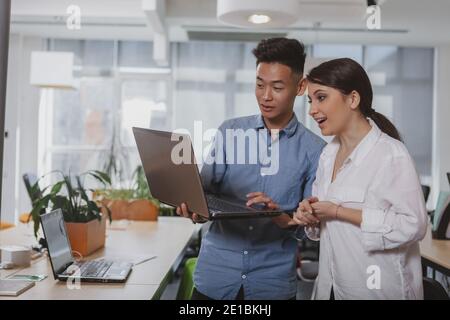 Asian businessman working on a laptop with his female colleague. Two young coworkers discussing startup ideas, browsing online on the computer, copy s Stock Photo