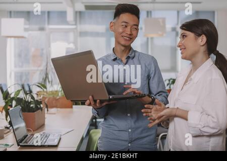 Young businesspeople laughing, talking at work, using laptop tigether. Asian businessman discussing ideas with his female colleague, copy space. Inter Stock Photo