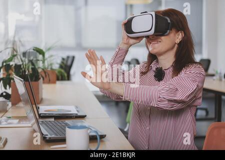 Cheerful mature woman smiling, weaeing 3d vr glasses at work. Happy businesswoman enjoying using virtual reality headset at the office. Future, techno Stock Photo