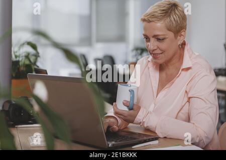 Lovely mature female entrepreneur working at her office, using laptop. Charming beautiful mature businesswoman enjoying working, having morning coffee Stock Photo