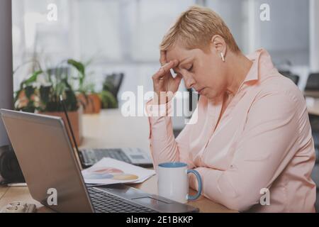 Mature businesswoman looking tired, rubbing her forehead, sitting in front of laptop. Experienced female entrepreneur having headache at work Stock Photo
