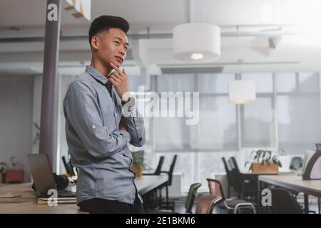 Thoughtful Asian businessman looking away, standing alone at his office, copy space. Confident male entrepreneur thinking about his project. Youth, ac Stock Photo