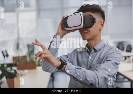 Cheerful Asian man smiling, using virtual reality 3d glasses, coy space. Happy male entrepreneur using 3d vr headset at his workplace. Cheerful busine Stock Photo