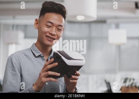 Close up of a happy young Asian man smiling, examining 3d vr glasses he is holding. Cheerful male entrepreneur preparing to use virtual reality headse Stock Photo