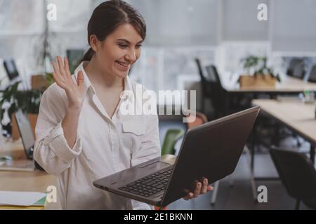 Cheerful young businesswoman smiling, waving at someone whi;le having video conference on her laptop. Lovely female entrepreneur video calling using h Stock Photo