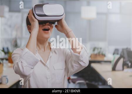 Excited young woman wearing 3d virtual reality goggles, copy space. Female entrepreneur using vr headset at work, looking surprised with her mouth ope Stock Photo