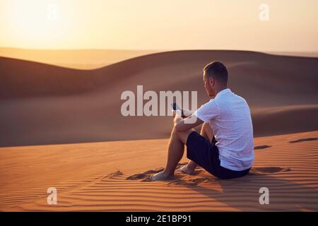 Alone in desert. Young man sitting on sand dune and using phone at golden sunset. Wahiba Sands in Oman. Stock Photo